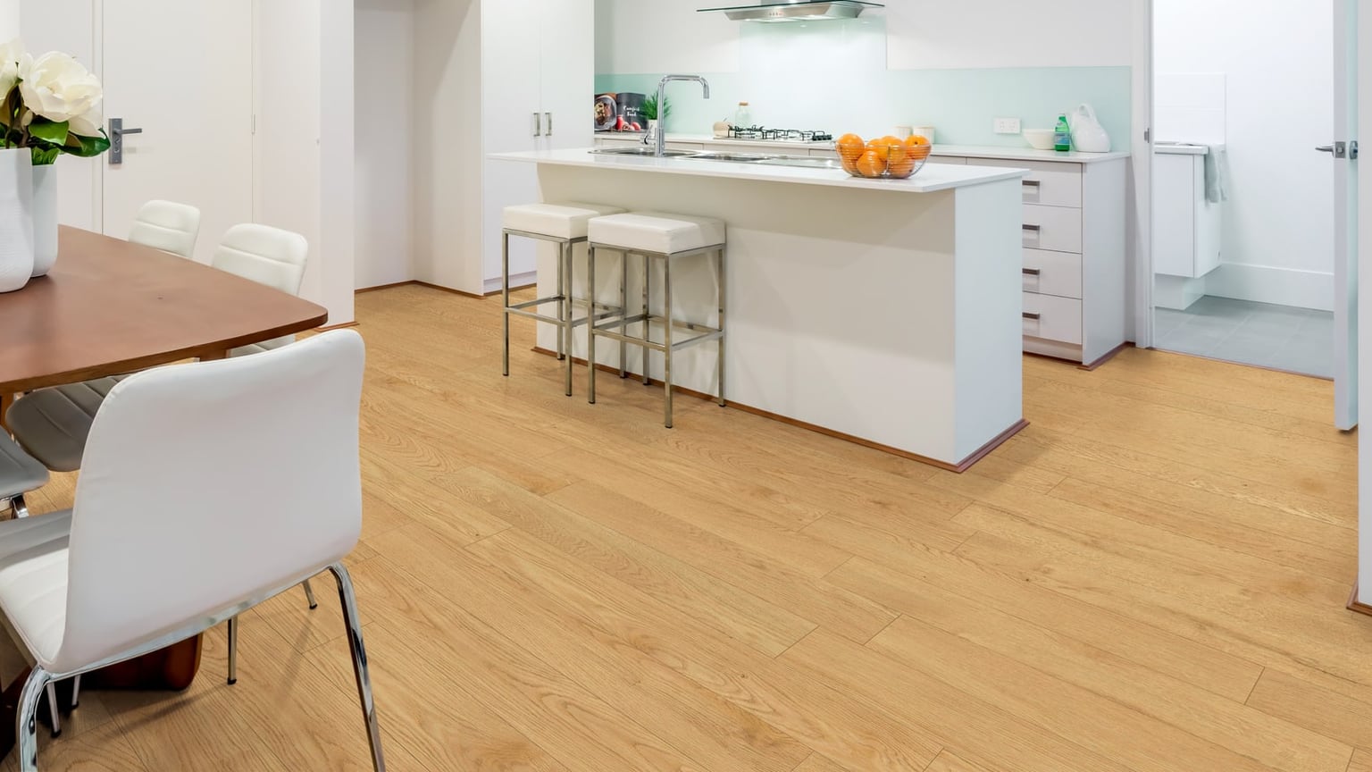 An open-plan dining area with a wooden table and white chairs beside a white kitchen island, over a light honey oak plank floor that fills the foreground.
