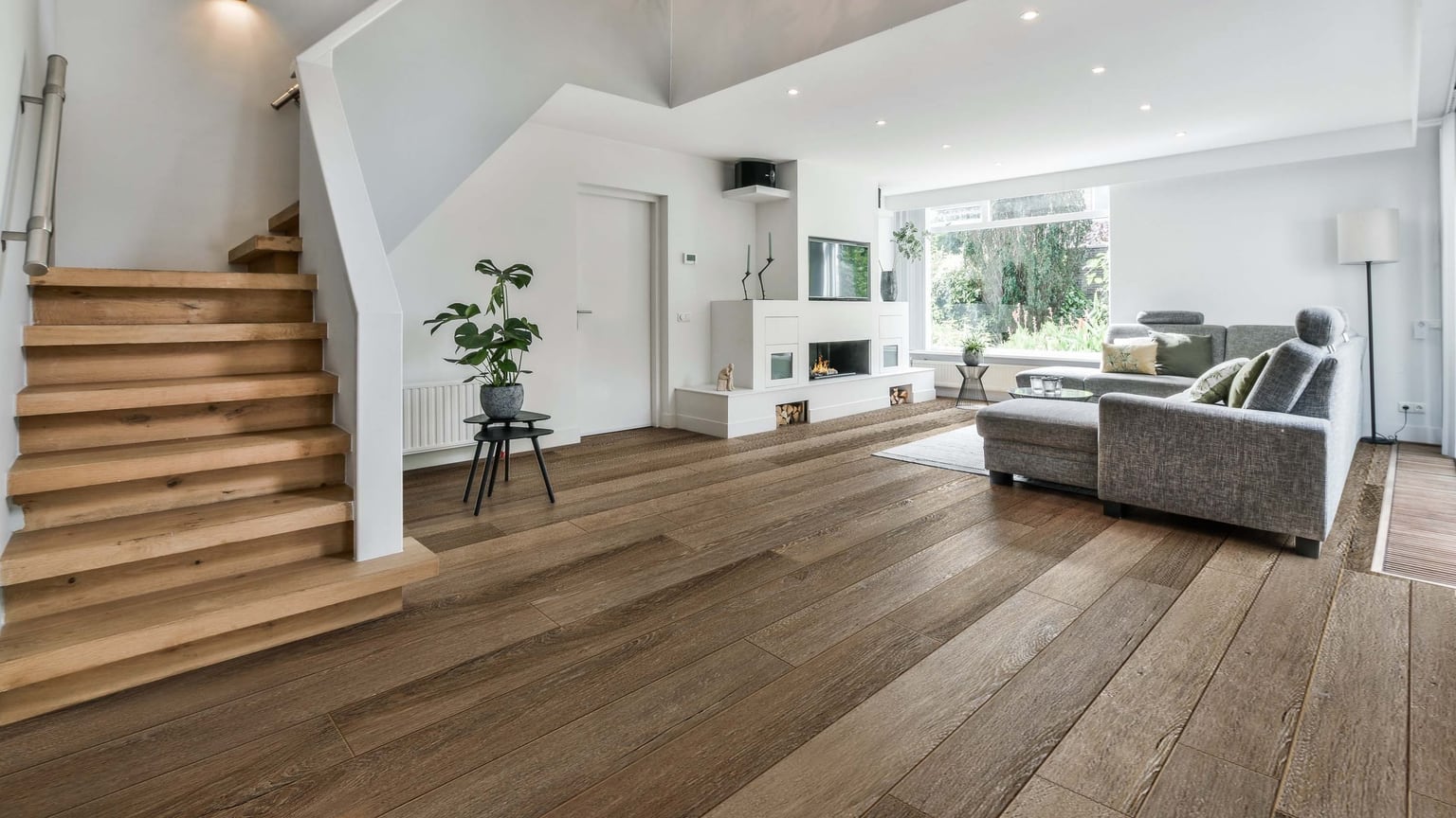 Open-plan living room with an open wooden staircase, white fireplace and grey corner sofa, with a warm brown plank floor filling the foreground.