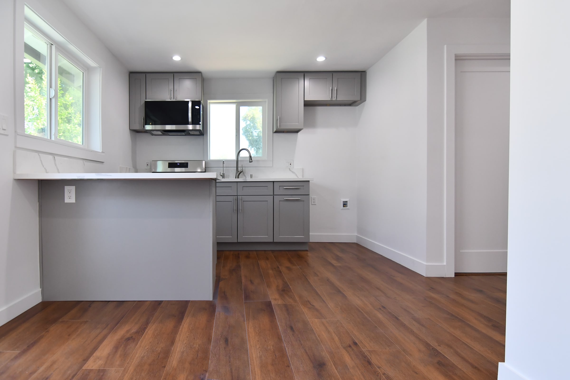 Empty kitchen with grey shaker cabinets, a white stone peninsula and a wide dark brown wood-look plank floor